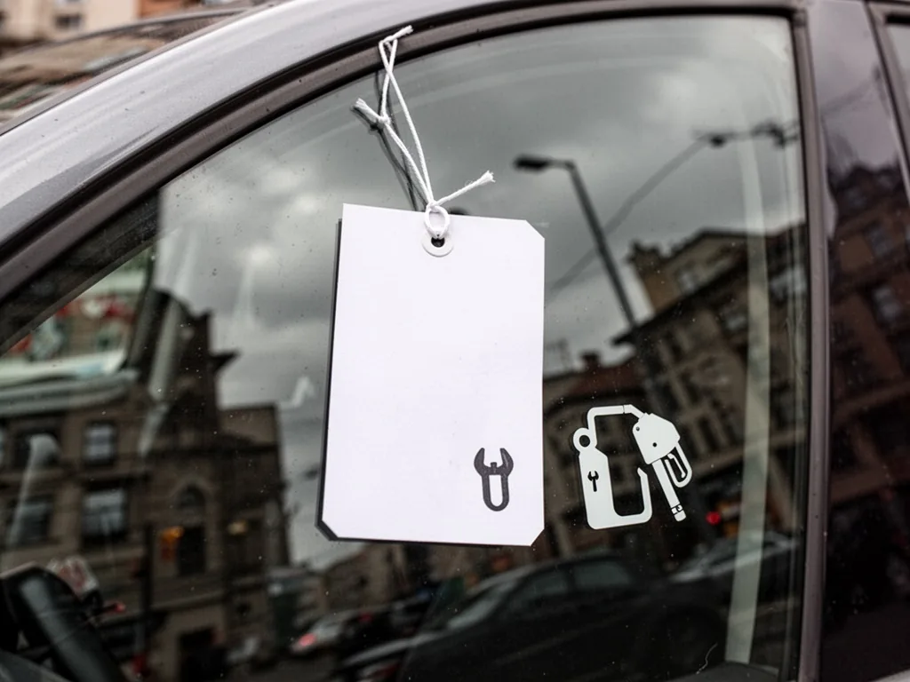 A white price tag attached to a car window, with a small wrench and fuel pump icon beside it, overcast city street light